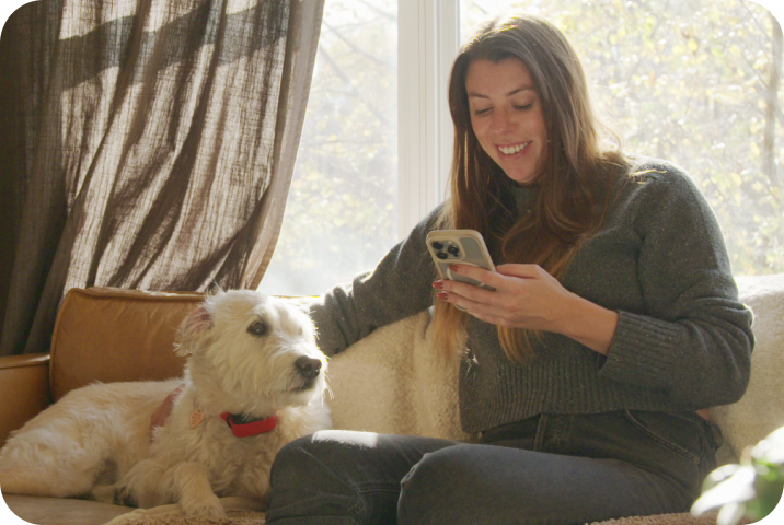 A woman sits on a couch looking at her phone while petting her dog.