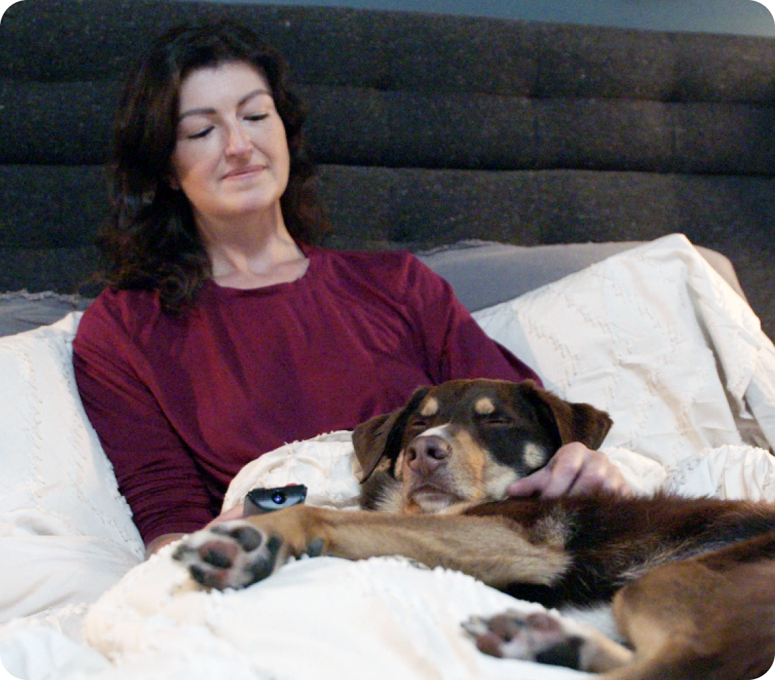 A woman sits in bed with her sleeping dog.