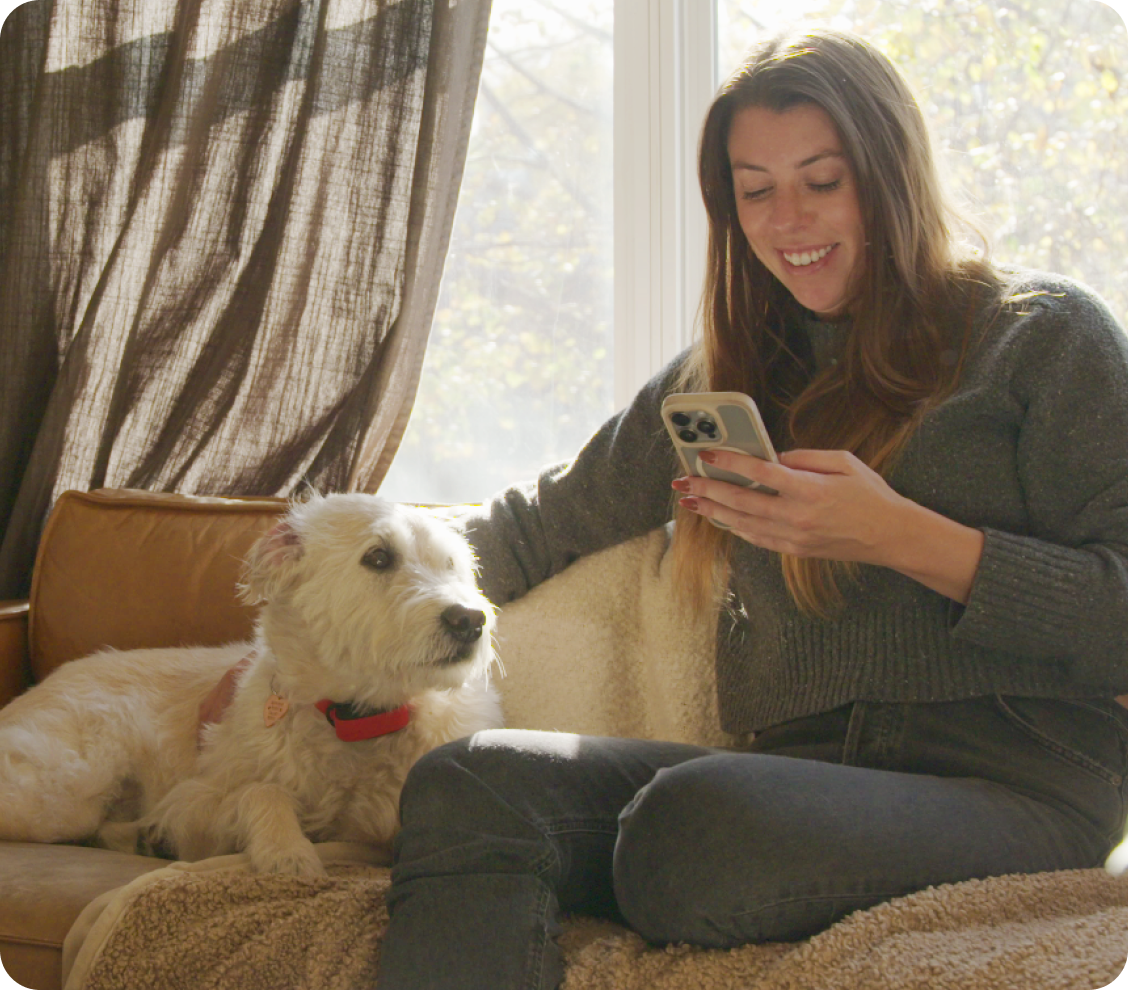 A woman sits on a couch looking at her phone while petting her dog.