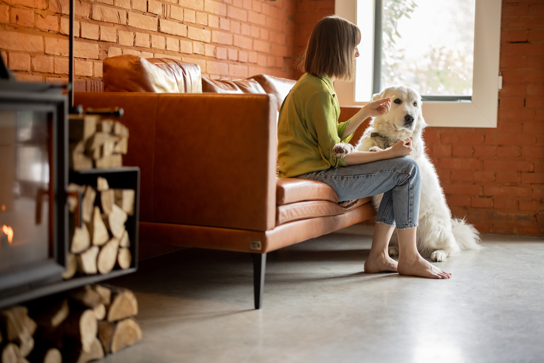 A woman sits on a couch holding a treat for her dog.